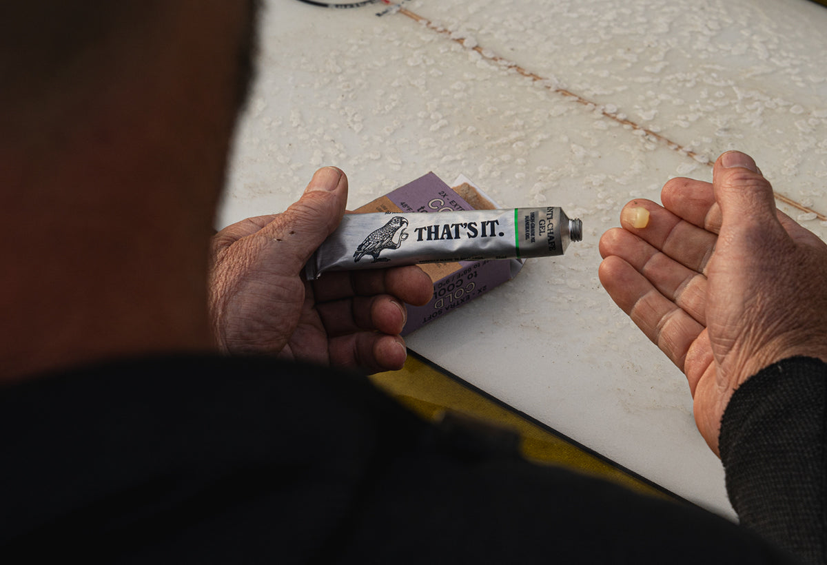 Person holding a tube of 'THAT'S IT.' product, leaning on a surfboard, applying to hand.